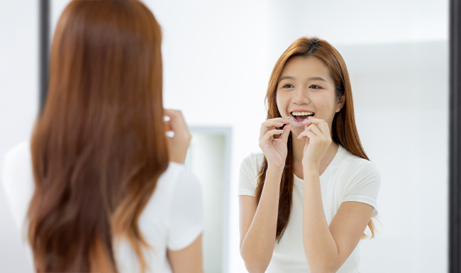 Young woman putting on retainer in front of mirror to maintain teeth alignment